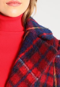 Abrigo de lana a cuadros con colores rojo, azul y amarillo; presenta una superficie suave y texturizada y un cuello de solapa, llevado sobre un suéter de cuello alto acanalado rojo.