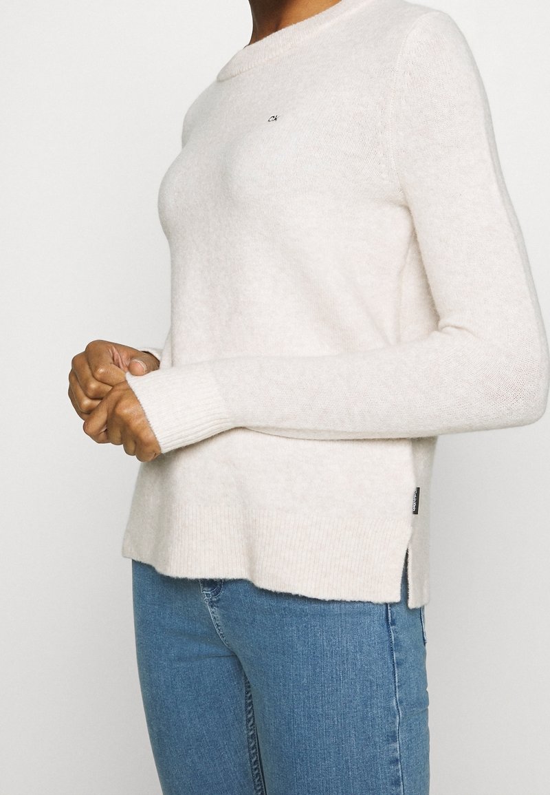 Personne portant un pull à manches longues de couleur crème avec un logo subtil et un jean bleu, debout les mains jointes devant.
