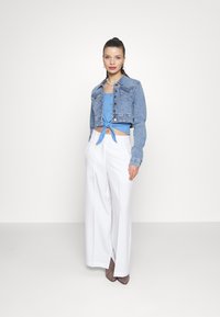 Giacca in denim cropped con chiusure a bottoni, top blu annodato e pantaloni bianchi a vita alta e gamba larga, abbinati a tacchi color taupe.
