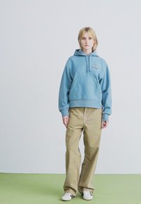 Una persona è in piedi su un pavimento verde contro una parete bianca semplice, indossando una felpa leggera blu, pantaloni beige a gamba larga e sneakers bianche.