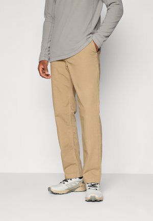 Person som har på seg beige bukser med rette bein, lys grå langermet skjorte og hvite joggesko med svarte lisser, står på en ensfarget bakgrunn.