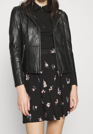 Nainen, joka pukeutunut mustaan nahkatakkiin mustan ribbineuleen ja mustan kukkakuosisen hameen päälle, jossa on vaaleanpunaisia ja sinisiä kukkia.