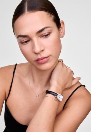 Montre carrée en argent avec un cadran blanc et un bracelet en cuir noir. Entourée de bijoux minimalistes, notamment un collier délicat en forme d'infini et des bagues.