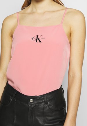 Kvinde iført en lyserød top med spaghetti-stropper med "CK Calvin Klein Jeans" logo og sorte læderbukser med høj talje.