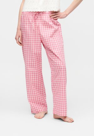 Κάτω μέρος σώματος ενός ατόμου που φοράει ροζ και άσπρα καρό φαρδιά πιτζαμαδάκια με σούρα στη μέση και γυμνά πόδια σε λευκό φόντο.