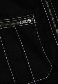 Svart denim tyg med vit sömnad, som har en dragkedjeficka och en strukturerad design. Dragkedjans metallkomponenter är i metall.