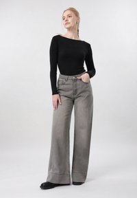 Ung kvinne med blondt hår i flette iført svart langermet topp og høytlivs grå vide jeans, stående med en hånd i lommen.