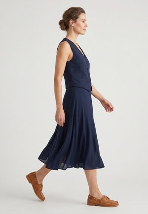 Kvinde går i navy ærmeløs knappet vest og matchende flydende midi nederdel med brune læder loafers på en ensfarvet baggrund.