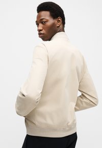 Jeune homme aux cheveux courts et bouclés portant une veste beige clair, légèrement tourné, regardant par-dessus son épaule sur un fond uni.