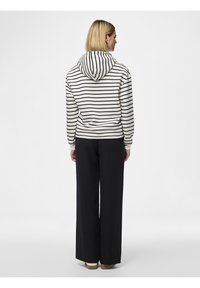 Kapuzensweatshirt in Creme mit schwarzen Streifen, featuring eine Fronttasche. Kombiniert mit lockeren schwarzen Hosen und hellen Schuhen.