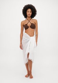 Hnědý bikiny top s křížovými ramínky, doplněný bílým texturovaným šátkem s bambulkami podél okraje. Modelka bosa.