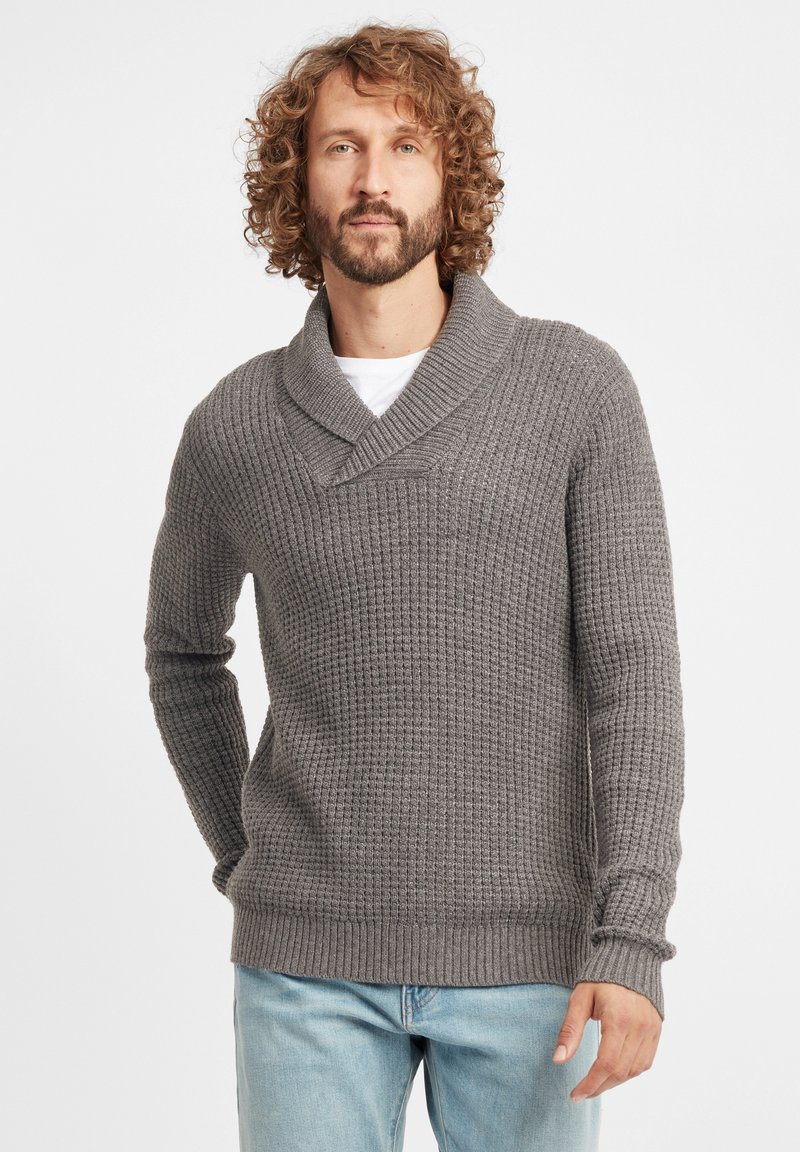 Grau gestrickter Pullover mit Schalkragen, der ein strukturiertes Muster und gerippte Bündchen aufweist. Getragen mit hellblauen Jeans.