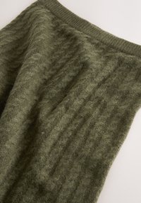 Jupe en laine tricotée douce de couleur vert olive avec un motif torsadé et une ceinture côtelée, posée à plat sur une surface claire.