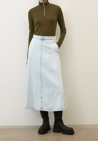 Olivengrünes Langarmshirt mit Reißverschluss, kombiniert mit einem hellblauen Jeans-Midrock mit Taschen und gerader Form, getragen mit schwarzen Stiefeln.