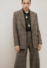 Kobieta w brązowej kratkowanej marynarce i spodniach o szerokich nogawkach, z warstwowymi złotymi naszyjnikami na czarnej bluzce, stojąca na jednolitym tle.