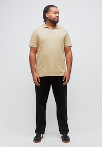 Άνδρας με πλεξούδες, φορώντας μπεζ πόλο πουκάμισο, μαύρο παντελόνι και μαύρα αθλητικά παπούτσια, στέκεται και κοιτάζει προς τα δεξιά σε απλό φόντο.