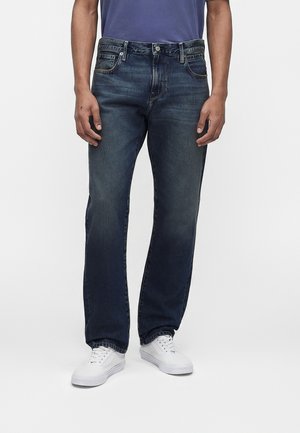 Mand iført mørkeblå jeans med lige ben og hvide sneakers, stående mod en ensfarvet lys baggrund.