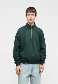 Tmavě zelená mikina s vysokým límcem a předním zipem. Měkká textura, volný střih a žebrované manžety se jednoduchým designem.