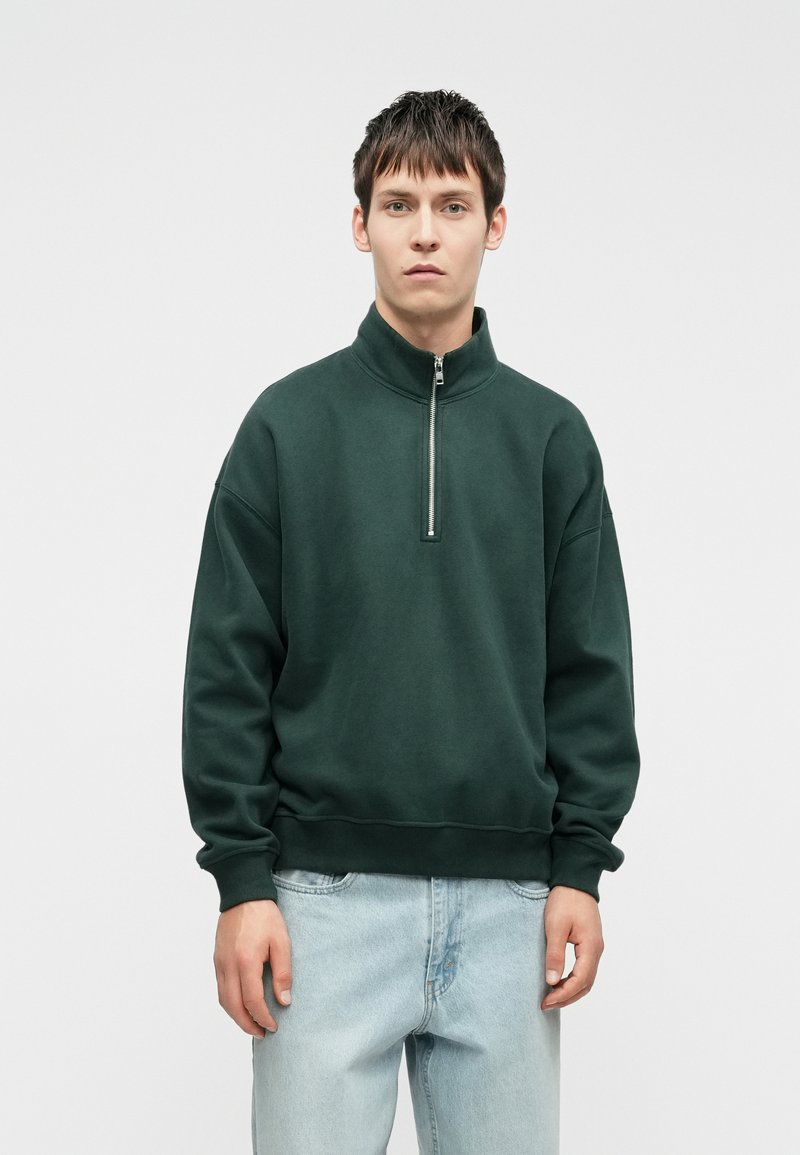 Tmavě zelená mikina s vysokým límcem a předním zipem. Měkká textura, volný střih a žebrované manžety se jednoduchým designem.