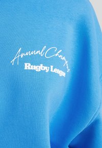 Sudadera azul de algodón con texto en blanco: "Annual Champions" en cursiva y "Rugby League" en una fuente en negrita. Textura lisa.