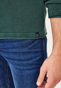 Grünes, geripptes Langarmhemd mit einem Etikett am Saum, kombiniert mit dunkelblauen Jeans. Nahaufnahme des unteren Saums und einer Hand, die auf der Jeans ruht.