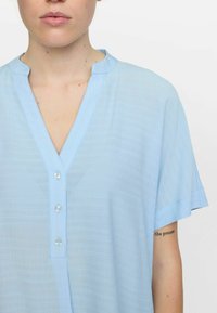 Lichtblauwe blouse met korte mouwen, gemaakt van textuurstof. Heeft een V-halslijn en vier knopen aan de voorkant voor sluiting.