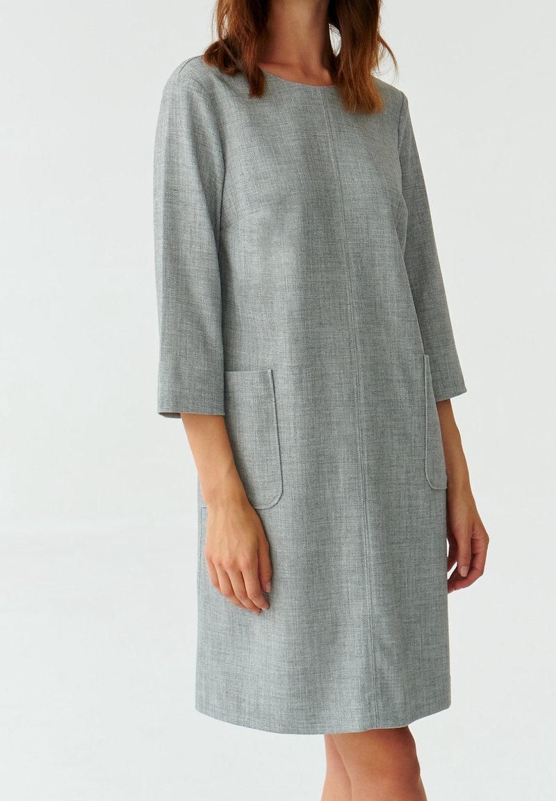 Donna che indossa un vestito grigio a lunghezza ginocchio con maniche tre quarti e tasche anteriori, in piedi davanti a uno sfondo bianco semplice.