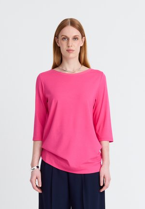 Woman with straight red hair wearing a bright pink three-quarter sleeve top and dark wide-leg pants against a plain light background.