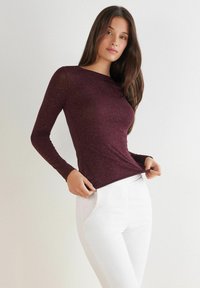 Frau mit langen braunen Haaren, die ein enges weinrotes Langarmoberteil und weiße Hosen trägt, posiert vor einem einfarbigen hellen Hintergrund.