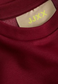 Burgundinės apvalios kaklų megztinis su raštuota apykakle, ant jo esantis audinio etiketė šviesiai smėlinė, ant kurios geltona spalva yra užrašas "JJXX". Minkšta tekstūra.
