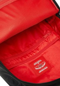 Interno di uno zaino nero con fodera rossa, dotato di una tasca con zip e un'etichetta in tessuto. Scomparti texturizzati e finitura superficiale liscia.