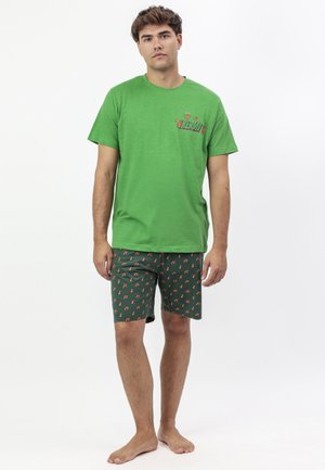 Joven de pie descalzo con una camiseta verde con un gráfico de Kermit y pantalones cortos oscuros con patrones de notas musicales naranjas.