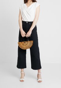 Top blanco drapeado, pantalones vaqueros de talle alto de pierna ancha en negro y tacones blancos con tiras. Sosteniendo un bolso de bambú en forma de media luna con un mango negro.