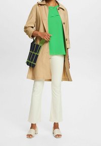 Trench coat khaki, top verde brillante, pantaloni bianchi a campana, mule con tacco quadrato color crema e borsa blu navy con motivo a griglia verde.