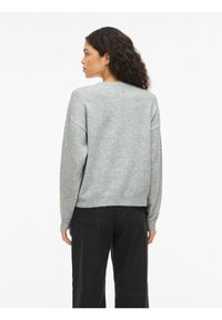 Grijze gebreide trui met afgezakte schouders, geribbelde zoom en een losse pasvorm, gecombineerd met zwarte broek, van achteren getoond. Gladde textuur.