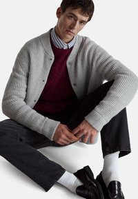 Graue, gerippte Strickjacke über einem dunkelroten Kabelstrickpullover, kombiniert mit dunklen Jeans und schwarzen Schuhen. Weiße Socken sind sichtbar.