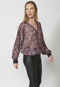Halbtransparente Bluse mit Paisleymuster in Lila- und Rosatönen, mit tiefem V-Ausschnitt und schwarzen, strukturierten Manschetten. Kombiniert mit schwarzen Hosen.