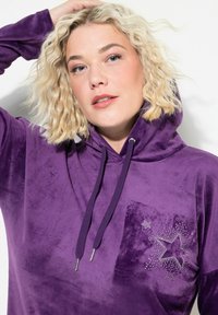 Vaalea nainen, jolla on kiharat hiukset, pukeutuneena purppuraan samettiin huppariin, jossa on rhinestone-tähtikuvio rinnassa, poseeraa toinen käsi pään takana.