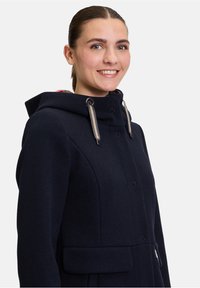 Marineblauer Wollmantel mit hohem Kragen und verstellbarem Kapuzenrand, ausgestattet mit silbernem Zubehör, versteckten Knöpfen und einem kontrastierenden karierten Futter.