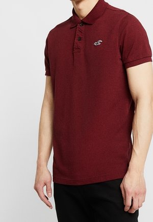 Man draagt een getailleerd bordeauxrood poloshirt met een klein wit logo op de linkerborst, gecombineerd met een zwarte broek tegen een effen achtergrond.