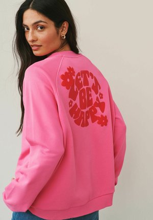 Young woman with long dark hair wears a pink sweatshirt with red "Let’s Be Happy" text and flower graphics on the back.