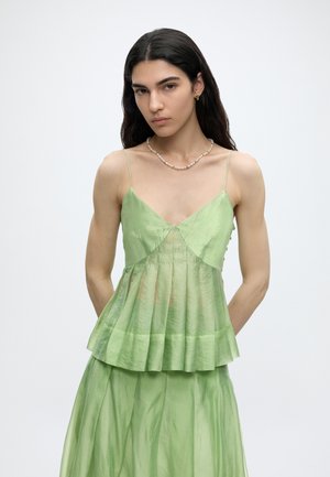 Žena s dlouhými tmavými vlasy, oblečená v světlém zeleném plisovaném topu bez rukávů a odpovídající sukni, stojící na bílém pozadí.