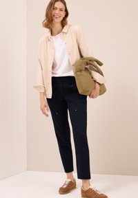 Donna sorridente, vestita con giacca beige chiaro, camicia bianca, pantaloni scuri, scarpe da ginnastica marroni, che tiene una borsa puffa verde oliva su uno sfondo beige.