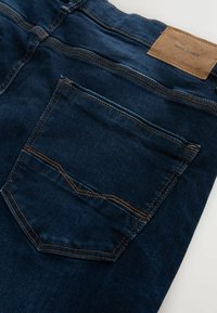 Tmavomodré džínsy vyrobené z denimu, které mají zadní kapsu s jemnými stehmi a koženou značku na pase. Hladká textura.