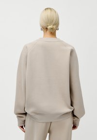 Person med blondt hår i en knute iført beige genser og matchende bukser, stående med ryggen til mot en ensfarget lys bakgrunn.