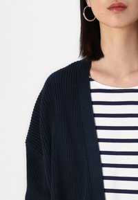 Granatowy sweter z dzianiny z teksturowanym wzorem i otwartym przodem, zestawiony z białą koszulką w granatowe paski pod spodem.