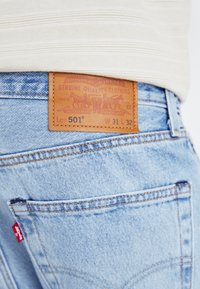 Ljusblå denimjeans med en brun läderetikett som har präglad design, storleksdetaljer och sömnadsdetaljer på bakfickan.