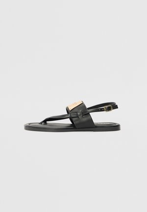 Black leather sandal with a flat sole, featuring a front thong design, adjustable ankle strap, and a matte gold metal accent.
