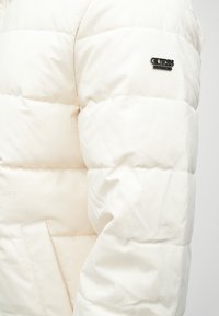 Πουκάμισο puffer σε απαλό λευκό με λεία υφή. Περιλαμβάνει ραμμένα τεμάχια, λογότυπο Guess και κρυφή μπροστινή τσέπη.