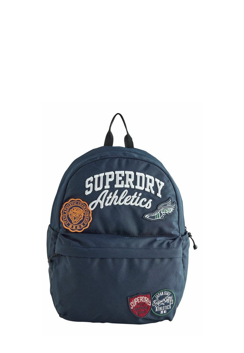 Zaino in tela navy con toppe ricamate, tra cui "SUPERDRY Athletics" e design circolari. Dotato di tasca con zip e manico superiore.